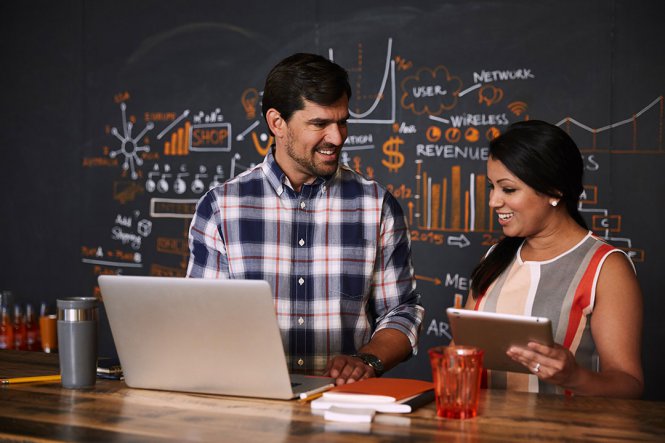 Smiling man and woman standing at laptops, in front of chalkboard.