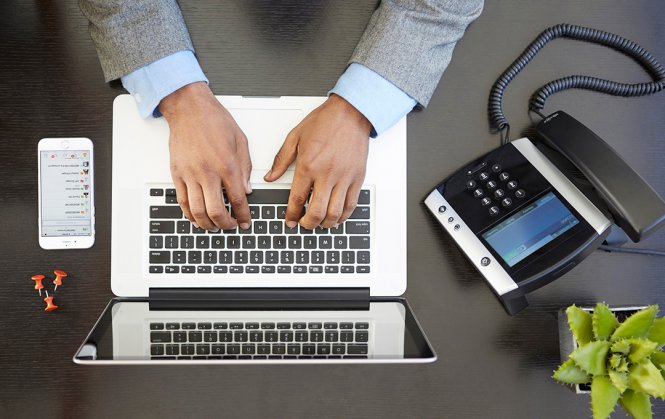Typing on a laptop, next to a smartphone and business phone.