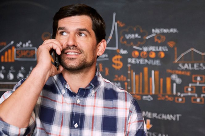 A man talking on a mobile phone, in front of a blackboard with figures on it.