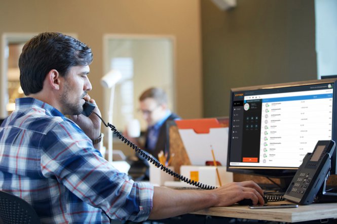 A man sitting at a computer, speaking on an office telephone.