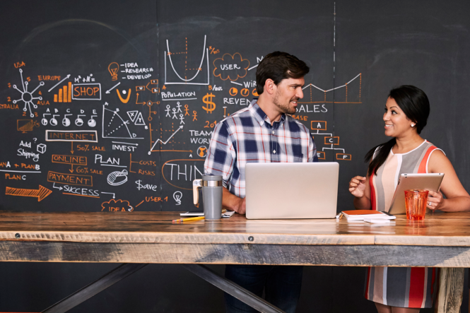 A man using a laptop, talking to a woman with a tablet.