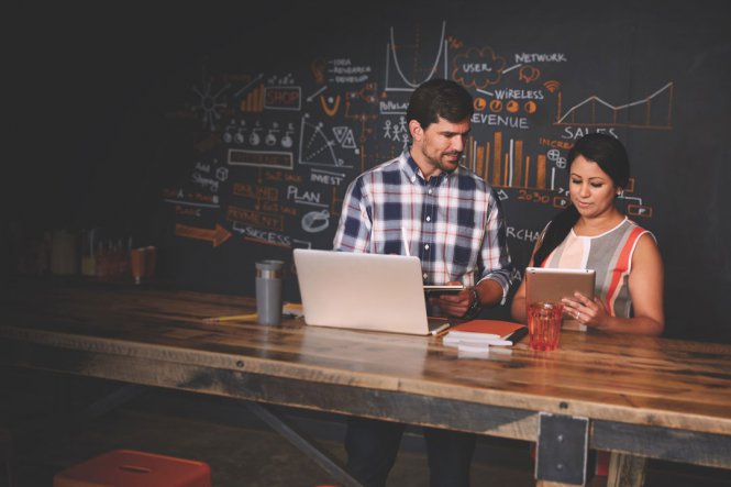 A man and a woman working on a laptop and tablet in front of a blackboard.