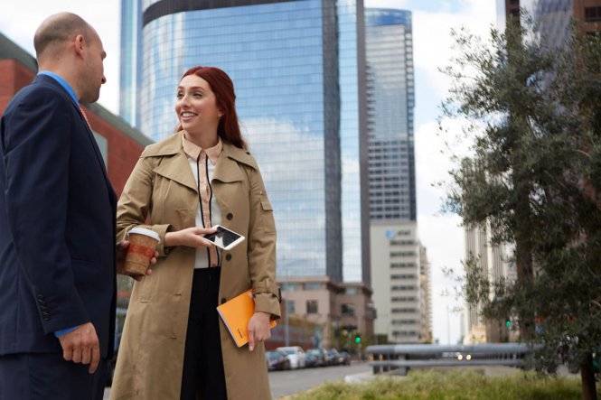 A woman holding a mobile phone is speaking to a man holding a cup of coffee, standing outside.