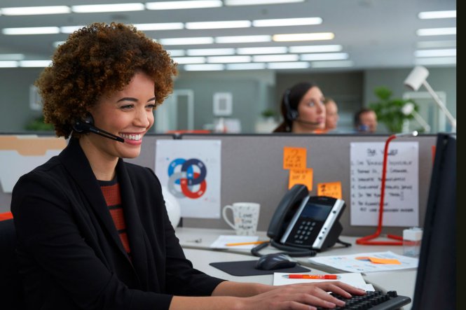 A woman sitting at an office computer, speaking on a headset.