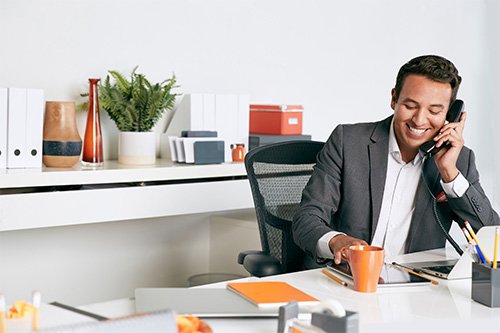 A smiling man talking on an office telephone while seated at a desk.