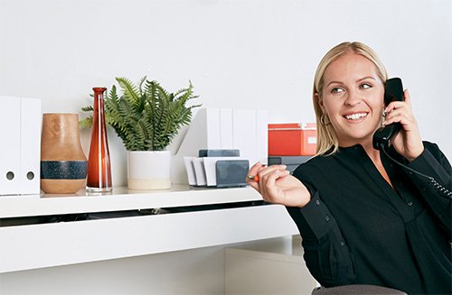 A smiling woman sitting at a desk, talking on an office telephone.