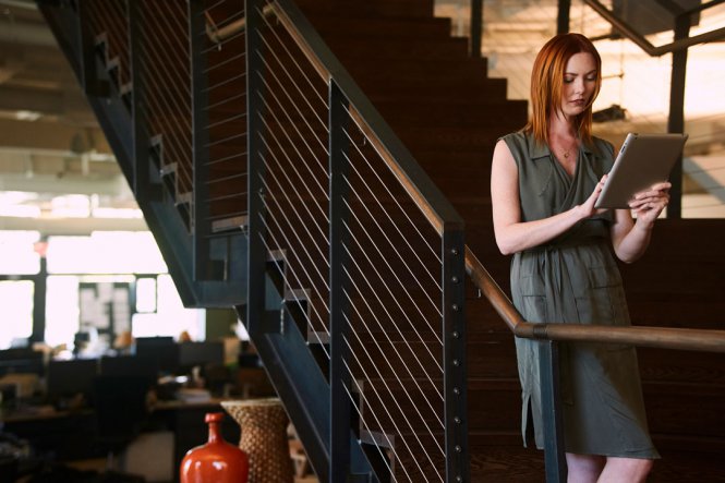 A woman is standing on the stairs in an office, using a tablet.