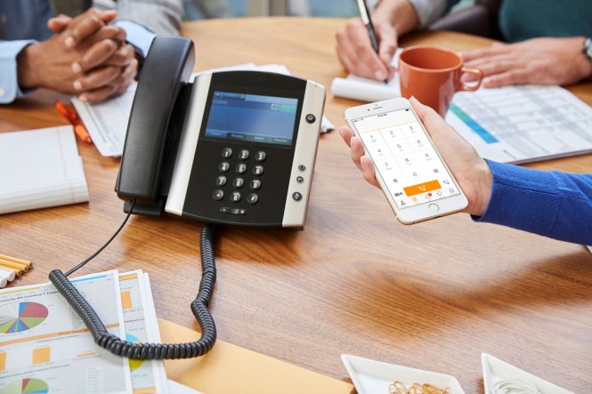 A close-up of an office telephone, and a person holding a smartphone next to it.