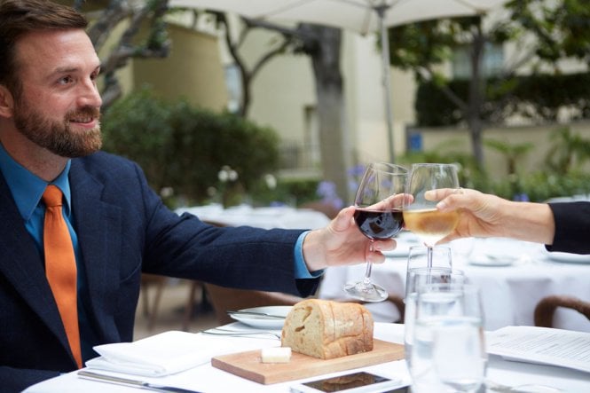 A man in a suit is sitting at a nice restaurant, enjoying wine with another person.
