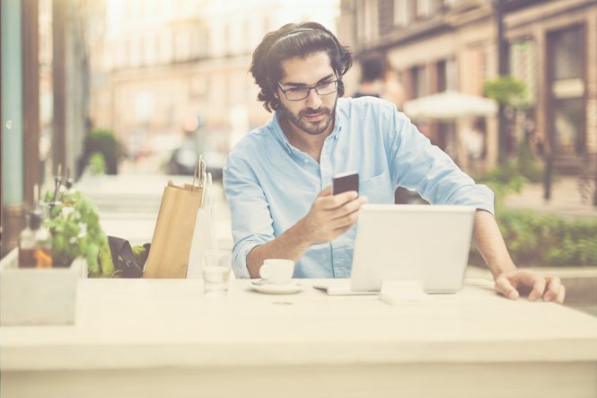 A man sitting outside at a cafe, using his mobile phone and laptop.
