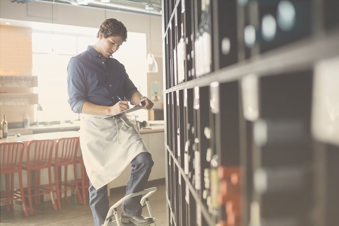 A man working at a restaurant, checking wine bottles and writing on a clipboard.