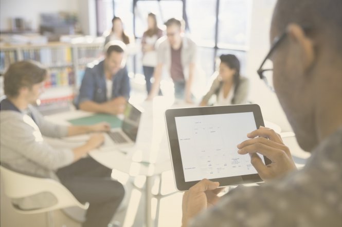 A man uses a tablet in an office, while his team members huddle around a table.