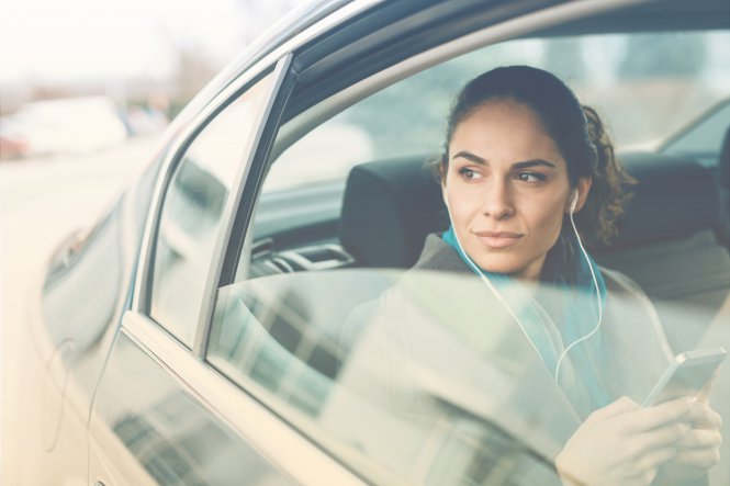 A woman using her smartphone with headphones while riding in the back seat of a car.