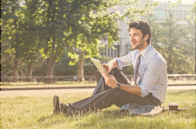 A man in a suit sitting in a park on a nice day, using a tablet.