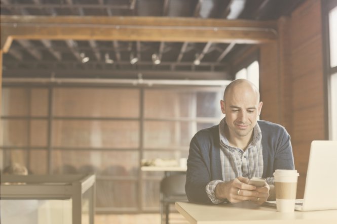 A man sitting in an open office, drinking coffee and using his smartphone.
