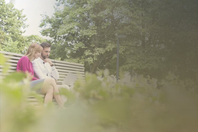 A man and a woman working on a laptop, sitting on a bench in a beautiful park.