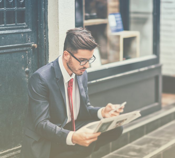 A man in a suit and using a mobile phone while reading a newspaper in the city.