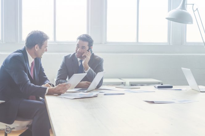 Two businessmen in a conference room with one enjoying software-defined WAN benefits while using his smartphone.