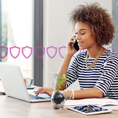 UC UCaaS Blog Header Photo of a woman in an office setting talking on her phone while typing or scrolling on her laptop. In the background, a series of small purple shields runs across the frame, representing security.