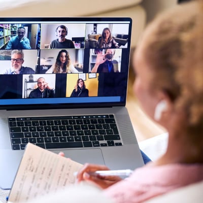 Image of a video conference meeting on a laptop with multiple participants in small video squares; in front of the laptop and blurred out is a woman with a notebook in her left hand and a pen in her right hand to take notes during the meeting
