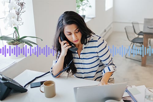 Photo of a woman in an office talking on her desk phone while typing on her computer. In the background, a series of small vertical purple-gradient lines run across the screen.