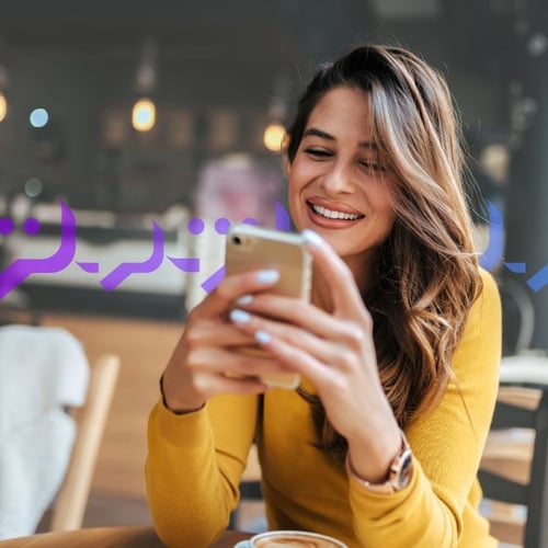 Photoillustration of a woman holding an iPhone in both of her hands and smiling at the screen; she has long, wavy brown hair and light coffee-colored skin and she's wearing coral lipstick; on top of the photo, there's an overlay graphic of message bubbles
