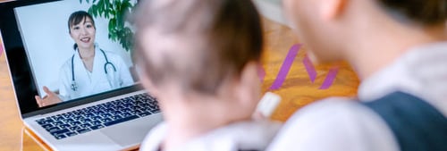Photo of a woman holding a baby in her arms with her back to the viewer; the woman and the baby are blurred out; the woman is facing an open laptop screen with a female, Asian American doctor on screen wearing a stethoscope, indicating a telehealth appointment 