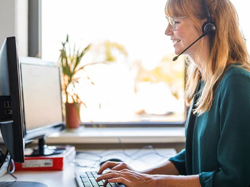 Photo of a smiling female call center agent talking to a customer on her headset as she types notes on her computer.