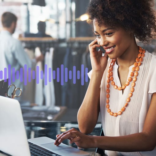 UC SEO Multichannel vs Omnichannel Blog Header Photo of a smiling employee in a store. She is talking on the phone while scrolling on her computer. In the background, a series of small purple vertical lines run across the frame, representing communication.