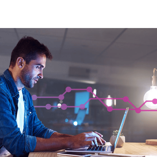 Photo of a man sitting at a table in an office setting typing on his laptop. Laying on the table in the foreground are a cell phone and a tablet. In the background, a series of purple lines and dots runs across the frame.
