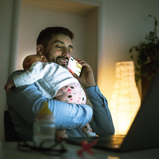 Photo of a business man at home, talking on his cellphone. He is holding a sleeping infant over his shoulder.