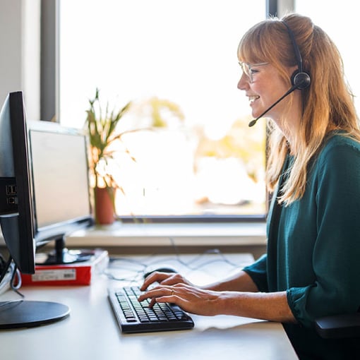 Photo of a smiling female call center agent talking to a customer on her headset as she types notes on her computer.