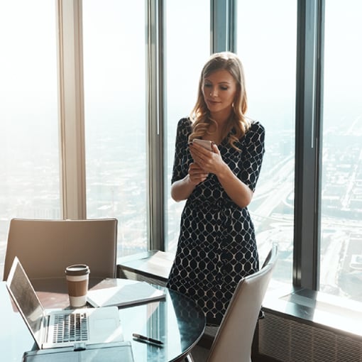 UC VoIP Cost For Business Thumbnail Photo of woman in an office. She is looking at something on her cellphone. On a table in the foreground are her laptop and some papers.