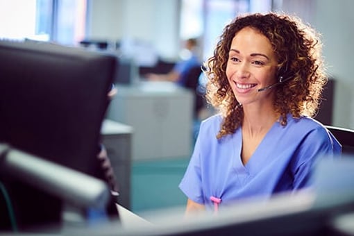 Image of healthcare worker wearing scrubs sitting in front of a computer with a headset on