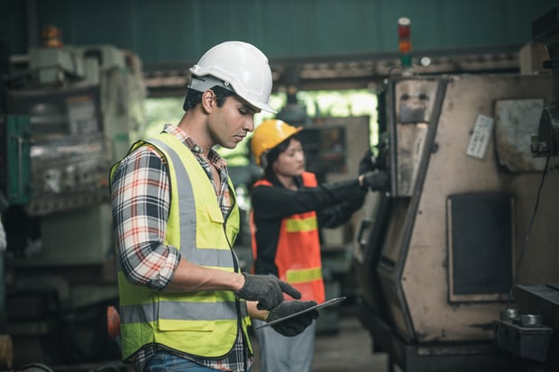 Getty image of two engineers wokring on heavy equipment in factory setting. The engineer in the foreground is checking specs on his tablet.
