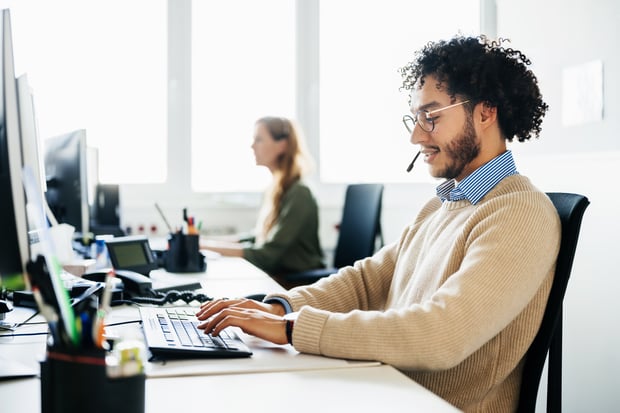 A man working at his computer at the office and talking to customers and clients on the phone using a hands free headset.