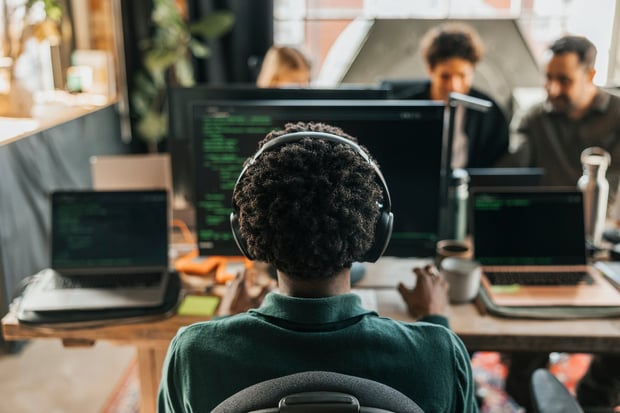 Rear view of curly haired male computer programmer wearing wireless headphones coding on desktop PC