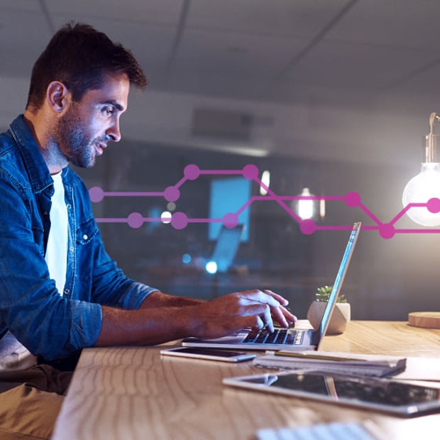 Photo of a man sitting at a table in an office setting typing on his laptop. Laying on the table in the foreground are a cell phone and a tablet. In the background, a series of purple lines and dots runs across the frame.