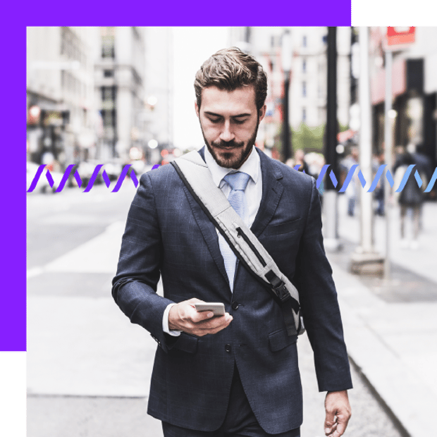 Photo of a man walking along a city street, wearing a suit with a backpack strap across his chest. He is looking down at a cell phone in his hand. In the background, a series of small wave forms runs across the frame.