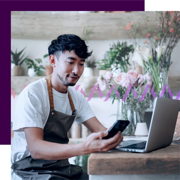 Photo of a male employee in a store with an open laptop on the counter, and he is looking at his cell phone.