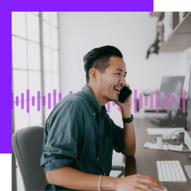 Photo of a smiling man in an office setting, talking on a cell phone and working on his computer.