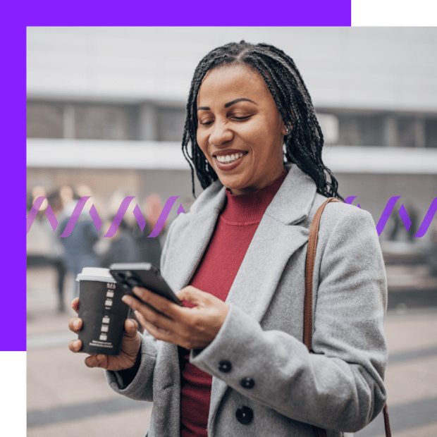 Photo of a smiling woman holding a coffee cup in one hand and her cell phone in the other. She is looking at the phone screen. She is outdoors in a city setting.