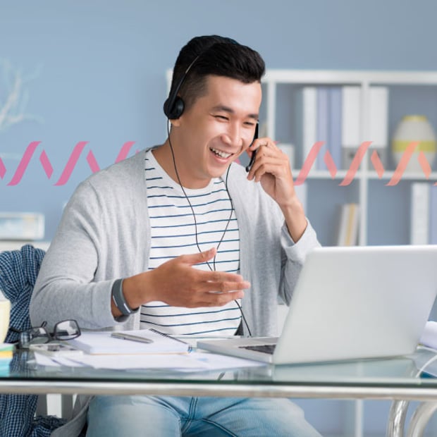 Forrester Whitepaper Blog Header Photo of a smiling employee working on his computer in an office. He is talking to someone on his phone and gesturing with his hand. In the background, a series of small gradient pink wave lines runs across the frame, representing communication