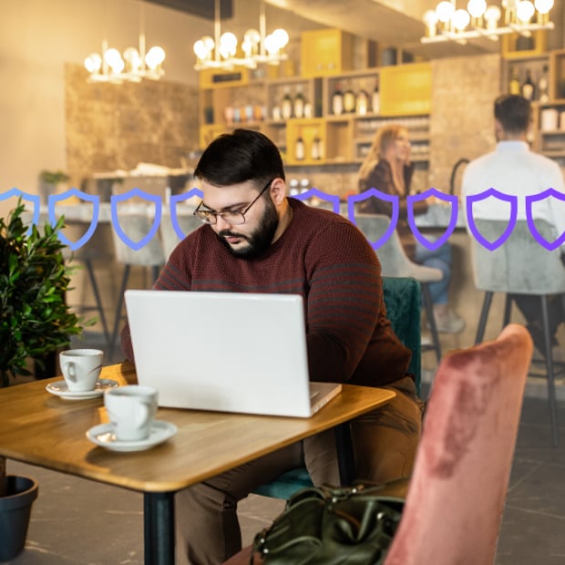 Network APIs SOC 2 Type 2 Blog Header Photo of a man sitting in a coffee shop working on his laptop. In the background, a series of small gradient-purple shield icons runs across the frame.