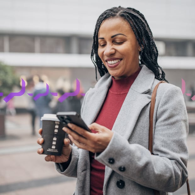 Traffic Control Blog Header Photo of a smiling woman outside of an office building, which is blurred in the background. She is looking at her phone in one hand and carrying a coffee cup in the other. Across the frame in the background run a series of small purple icons representing text traffic.