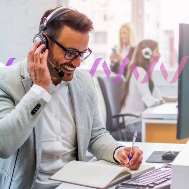 Photo of a smiling male call center agent talking to a customer through his headset and taking notes 