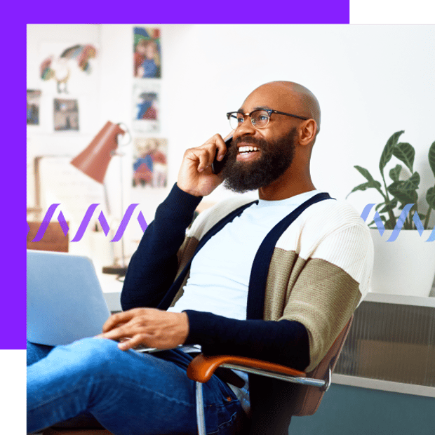Photo of a smiling man sitting in an office with his laptop on his lap. He is talking on his mobile phone. In the background, a series of small purple wave forms run across the frame.