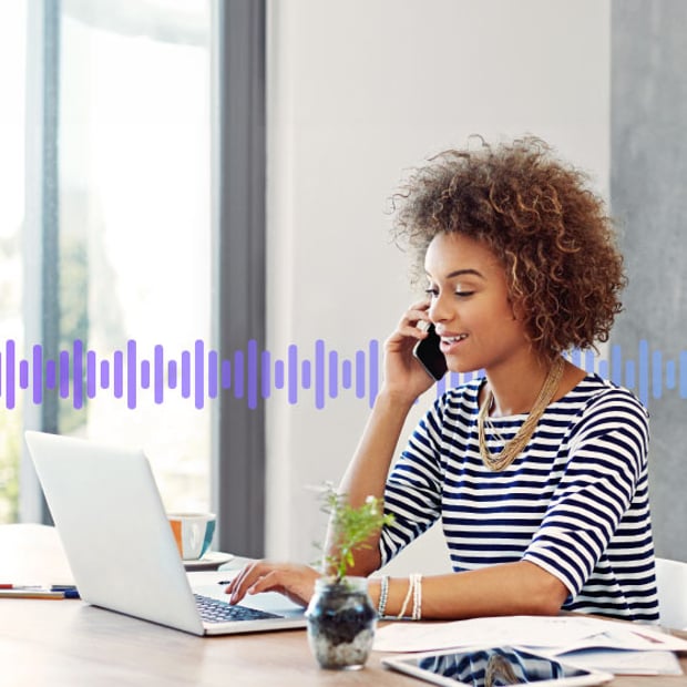 UC SEO How to Forward Voicemail Blog Header Photo of a woman in an office talking on her phone while typing on her laptop. Running across the frame in the background are a series of small purple vertical lines representing a voicemail recording.