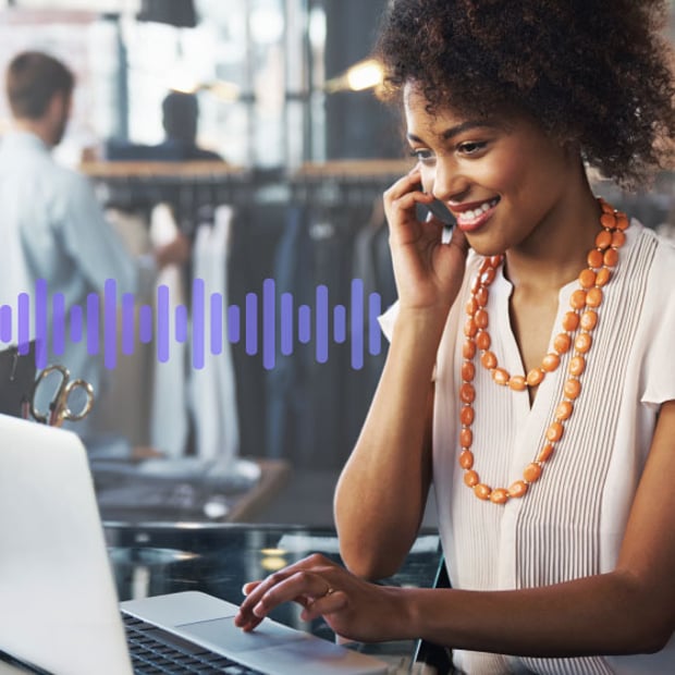 UC SEO Multichannel vs Omnichannel Blog Header Photo of a smiling employee in a store. She is talking on the phone while scrolling on her computer. In the background, a series of small purple vertical lines run across the frame, representing communication.