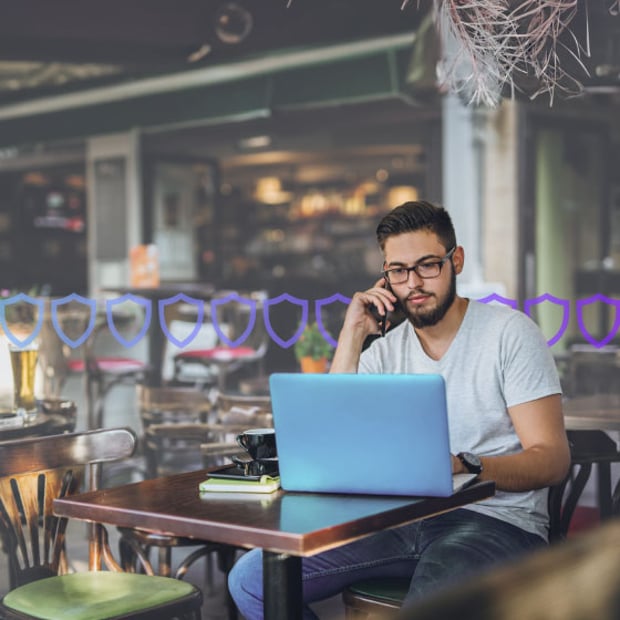 UC SEO VoIP Security Blog Header Photo of a casually dressed man sitting alone at a table in a restaurant working on his laptop and talking on his cell phone. Running across the frame are a series of small shield icons representing security.